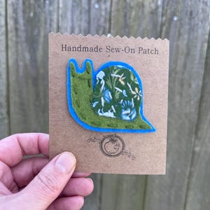 May include: A handmade sew-on patch featuring a green snail with a blue outline. The snail's shell is patterned with a floral design in shades of green and white. The patch is on a brown card with the text "Handmade Sew-On Patch".