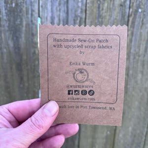 Puede incluir: Una etiqueta de papel marr&oacute;n con el texto "Handmade Sew-On Patch with upcycled scrap fabrics by Erika Wurm." La etiqueta tambi&eacute;n incluye iconos de redes sociales y el sitio web erikawurm.com.