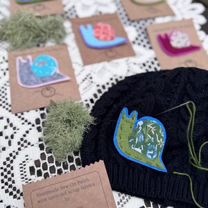 Puede incluir: Parches cosidos a mano con dise&ntilde;os de caracoles coloridos. Los parches est&aacute;n hechos con retales de tela reciclados. La imagen muestra varios parches sobre tarjetas marrones, con un parche de caracol verde y azul cosido a un gorro negro. El texto de la tarjeta dice "Erika Wurm".