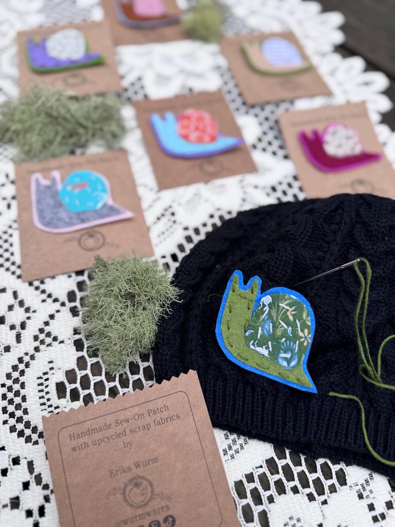Puede incluir: Parches cosidos a mano con dise&ntilde;os de caracoles coloridos. Los parches est&aacute;n hechos con retales de tela reciclados. La imagen muestra varios parches sobre tarjetas marrones, con un parche de caracol verde y azul cosido a un gorro negro. El texto de la tarjeta dice "Erika Wurm".