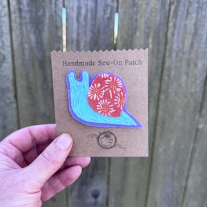 May include: A handmade sew-on patch featuring a snail design. The snail has a light blue body, a red shell with white and orange floral patterns, and a purple outline. The patch is on a brown card with the text "Handmade Sew-On Patch".