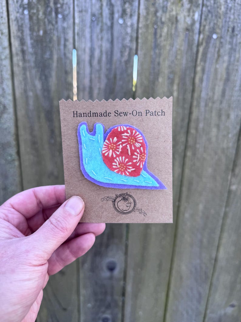 Puede incluir: Un parche cosido a mano con un dise&ntilde;o de caracol. El caracol tiene un cuerpo azul claro, una concha roja con patrones florales blancos y naranjas, y un contorno morado. El parche est&aacute; en una tarjeta marr&oacute;n con el texto "Handmade Sew-On Patch".