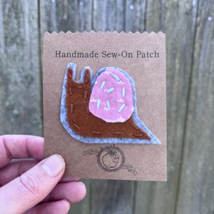 May include: A handmade sew-on patch featuring a brown snail with a pink shell decorated with blue sprinkles. The snail is outlined in gray and stitched with brown thread. The patch is on a brown card with the text "Handmade Sew-On Patch".