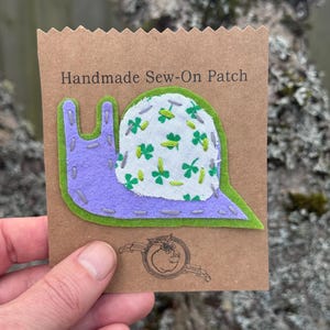 May include: A handmade sew-on patch featuring a whimsical snail design. The snail has a purple body, a white shell adorned with green shamrocks, and a green border. The patch is on a brown card with text.