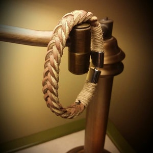 May include: A bracelet featuring a braided tan leather strap connected to a section of wrapped beige twine. The ends of the bracelet are secured with polished silver-toned metal cylindrical clasps that meet to form a circular closure.