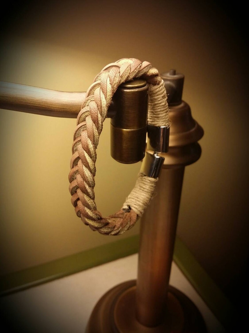 May include: A bracelet featuring a braided tan leather strap connected to a section of wrapped beige twine. The ends of the bracelet are secured with polished silver-toned metal cylindrical clasps that meet to form a circular closure.