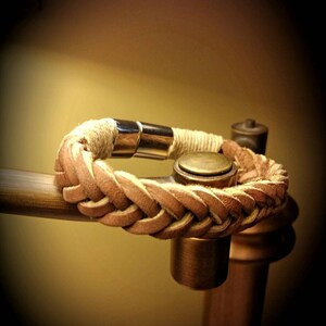 May include: A braided tan leather bracelet featuring a polished silver-tone metal cylindrical clasp. The leather strands are woven into a thick, textured band, with a small section of light-coloured cord wrapping near the metallic closure.