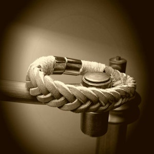 May include: A braided tan leather bracelet featuring a polished silver-tone metal cylindrical clasp. The bracelet is constructed from multiple strands of leather woven into a thick, textured band, secured by a smooth, reflective metal connector.