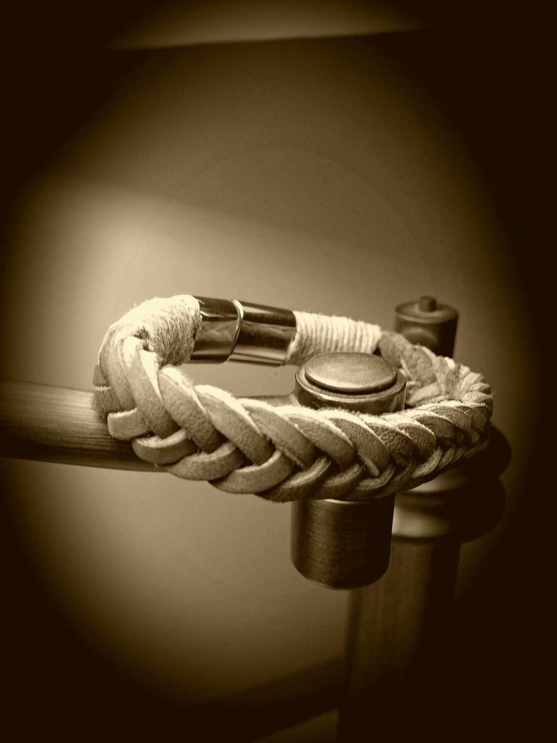 May include: A braided tan leather bracelet featuring a polished silver-tone metal cylindrical clasp. The bracelet is constructed from multiple strands of leather woven into a thick, textured band, secured by a smooth, reflective metal connector.