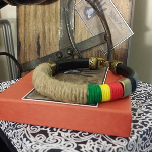 May include: A handmade bracelet with a hemp rope design, accented with green, yellow, and red bands. The bracelet features brass-colored metal clasps and accents. It rests on a red box atop a patterned cloth.