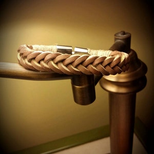 May include: A braided tan leather bracelet featuring a smooth, light-coloured cord wrapping near the ends and a polished silver-toned metal cylindrical clasp. The leather strands are woven in a consistent pattern, creating a textured appearance.