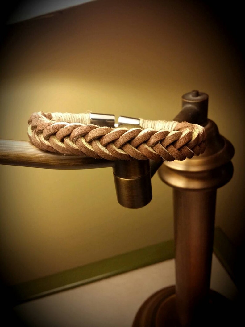 May include: A braided tan leather bracelet featuring a smooth, light-coloured cord wrapping near the ends and a polished silver-toned metal cylindrical clasp. The leather strands are woven in a consistent pattern, creating a textured appearance.