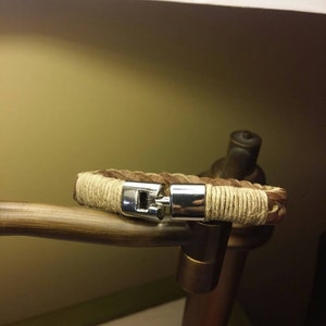 May include: A braided brown leather bracelet featuring sections wrapped in light tan twine and a polished silver-tone metal clasp. The clasp mechanism is centred, connecting the two ends of the leather and twine-wrapped band securely.
