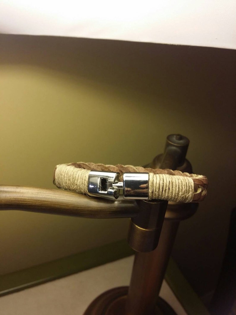 May include: A braided brown leather bracelet featuring sections wrapped in light tan twine and a polished silver-tone metal clasp. The clasp mechanism is centred, connecting the two ends of the leather and twine-wrapped band securely.