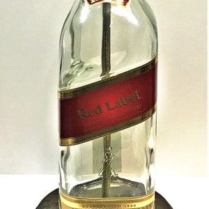 Puede incluir: Una l&aacute;mpara de mesa hecha de una botella de vidrio transparente con una etiqueta dorada que dice "Johnnie Walker Red Label Blended Scotch Whisky Established 1820 1.75 Litre 40% Alc./Vol.  Imported by Diageo North America, CT". La botella est&aacute; sobre una base de madera marr&oacute;n oscuro.