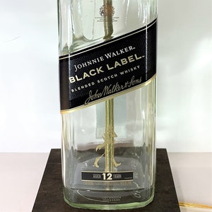 May include: A clear glass bottle lamp with a gold metal base and a black label that reads "Johnnie Walker Black Label Blended Scotch Whisky". The bottle is sitting on a dark brown wooden base.
