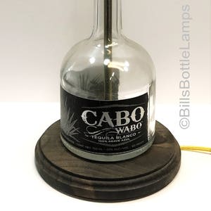 May include: A clear glass bottle lamp with a black label that reads "CABO WABO TEQUILA BLANCO". The lamp has a gold-colored socket and sits on a dark wooden base. The lamp is approximately 12 inches tall.