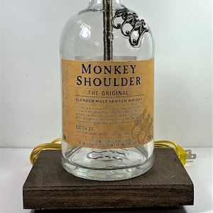 May include: A clear glass bottle lamp with a gold metal lampshade and a metal monkey shoulder design. The bottle is labeled "Monkey Shoulder The Original Blended Malt Scotch Whisky" and "Batch 27".