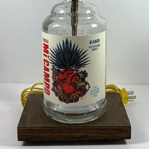 May include: A repurposed tequila bottle lamp with a brass lamp holder. The clear glass bottle features a colorful label with a heart and agave design. The lamp sits on a dark wooden base and has a yellow cord.