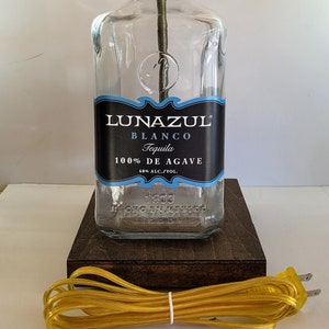 May include: A clear glass bottle lamp with a gold metal lampshade. The bottle is labeled "Lunazul Blanco Tequila 100% de Agave 40% Alc./Vol.". The lamp is sitting on a dark brown wooden base and has a yellow cord.