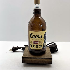 May include: A brown glass beer bottle with a Coors Golden Banquet Beer label. The bottle is turned into a lamp with a brown cardboard lampshade and a brown wooden base.