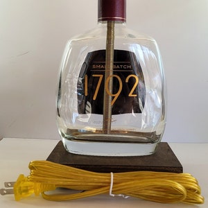 May include: A clear glass bottle lamp with a gold colored lampshade and a brown wooden base. The bottle has a label that says "Small Batch 1792" and a yellow electrical cord.
