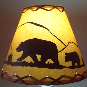 May include: A brown lampshade with a silhouette of two bears walking in a mountain landscape. The lampshade has a brown leather trim with stitching.