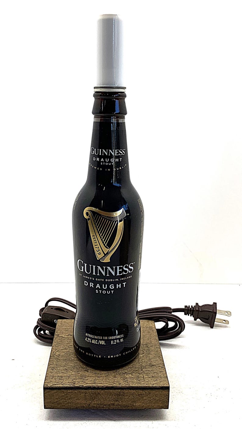 May include: A Guinness Draught Stout bottle converted into a lamp. The black bottle features the Guinness logo and text, with a white lamp shade and a wooden base. The lamp has a brown cord and a standard US plug.