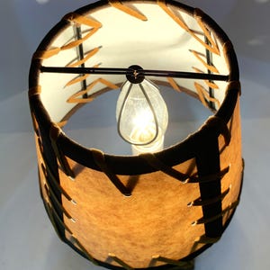 May include: A small, illuminated lamp with a cylindrical lampshade. The shade is brown with a stitched design and a dark brown trim. The interior is white, and a light bulb is visible inside. The lamp is on a white surface.