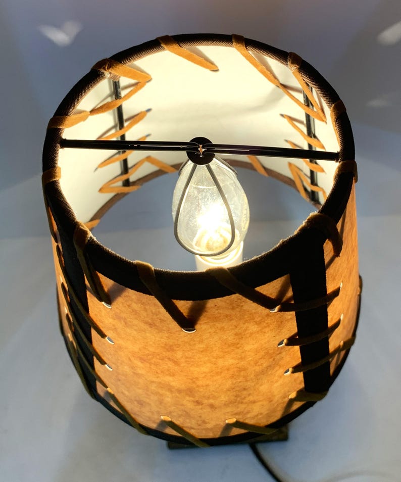 May include: A small, illuminated lamp with a cylindrical lampshade. The shade is brown with a stitched design and a dark brown trim. The interior is white, and a light bulb is visible inside. The lamp is on a white surface.
