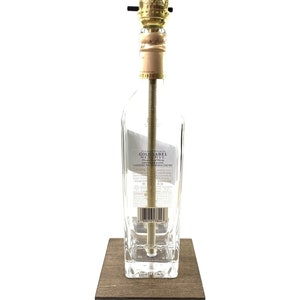 May include: A repurposed clear glass bottle lamp with a gold metal lampshade and a wooden base. The bottle label reads "Johnnie Walker Gold Label Reserve Blended Scotch Whisky".