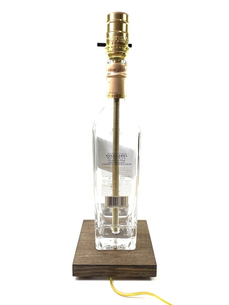 May include: A repurposed clear glass bottle lamp with a gold metal lampshade and a wooden base. The bottle label reads "Johnnie Walker Gold Label Reserve Blended Scotch Whisky".