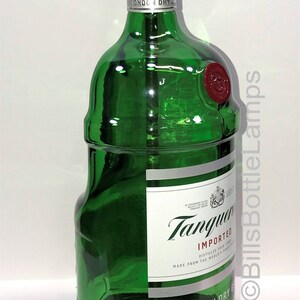 Puede incluir: Una l&aacute;mpara de botella de vidrio verde con una base de madera. La botella est&aacute; etiquetada con "Tanqueray Imported London Dry Gin" y tiene un sello de cera rojo. La l&aacute;mpara est&aacute; encendida con una bombilla dorada.