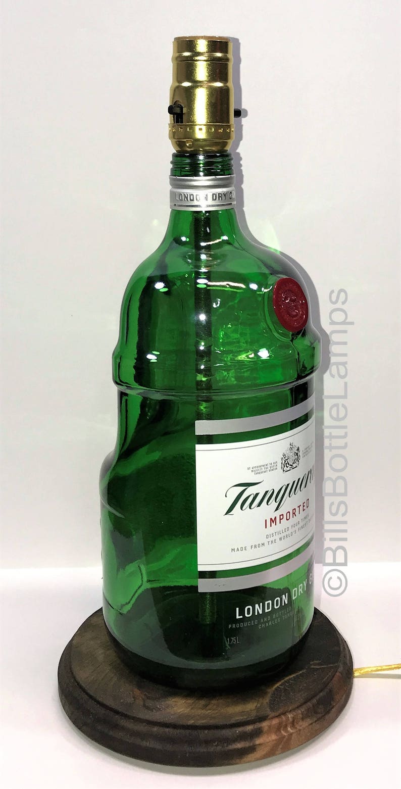 Puede incluir: Una l&aacute;mpara de botella de vidrio verde con una base de madera. La botella est&aacute; etiquetada con "Tanqueray Imported London Dry Gin" y tiene un sello de cera rojo. La l&aacute;mpara est&aacute; encendida con una bombilla dorada.