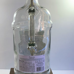 May include: A clear glass bottle lamp with a gold metal lampshade and a brown wooden base. The bottle has a label that reads "Tanqueray Rangpur" and includes serving facts and nutritional information. The lamp is plugged in with a yellow cord.