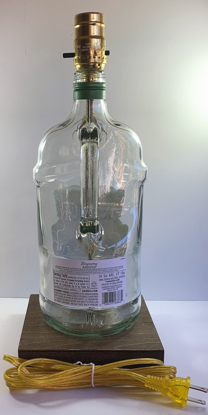 May include: A clear glass bottle lamp with a gold metal lampshade and a brown wooden base. The bottle has a label that reads "Tanqueray Rangpur" and includes serving facts and nutritional information. The lamp is plugged in with a yellow cord.