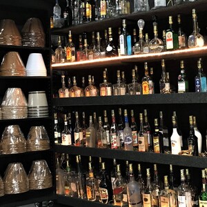 Op de afbeelding: Een muur met planken vol glazen flessen, elk met een gouden dop. De flessen zijn gerangschikt in rijen en kolommen, en ze zijn allemaal van verschillende grootte en vorm. Er zijn ook verschillende stapels lampenkappen op de foto, die bruin en wit zijn.