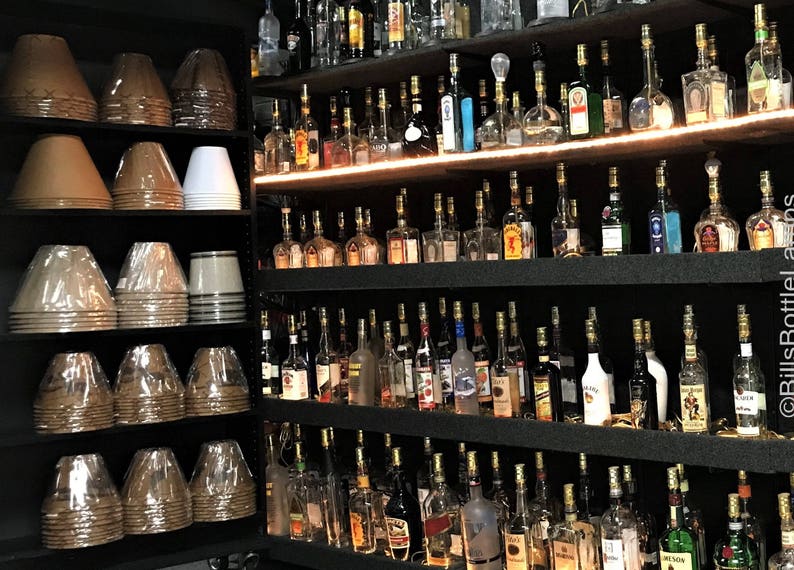 Op de afbeelding: Een muur met planken vol glazen flessen, elk met een gouden dop. De flessen zijn gerangschikt in rijen en kolommen, en ze zijn allemaal van verschillende grootte en vorm. Er zijn ook verschillende stapels lampenkappen op de foto, die bruin en wit zijn.