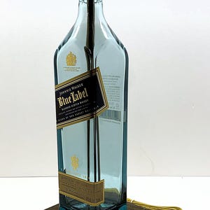 K&ouml;nnte beinhalten: Eine umfunktionierte Lampe aus einer Johnnie Walker Blue Label Whiskyflasche. Die blau get&ouml;nte Glasflasche hat ein schwarz-goldenes Etikett. Die Lampe hat eine goldfarbene Fassung und steht auf einem dunklen Holzsockel.