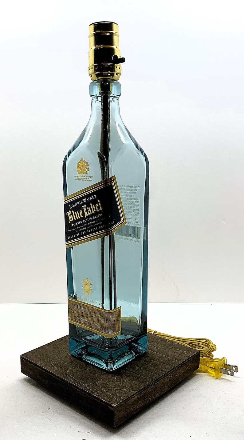 K&ouml;nnte beinhalten: Eine umfunktionierte Lampe aus einer Johnnie Walker Blue Label Whiskyflasche. Die blau get&ouml;nte Glasflasche hat ein schwarz-goldenes Etikett. Die Lampe hat eine goldfarbene Fassung und steht auf einem dunklen Holzsockel.