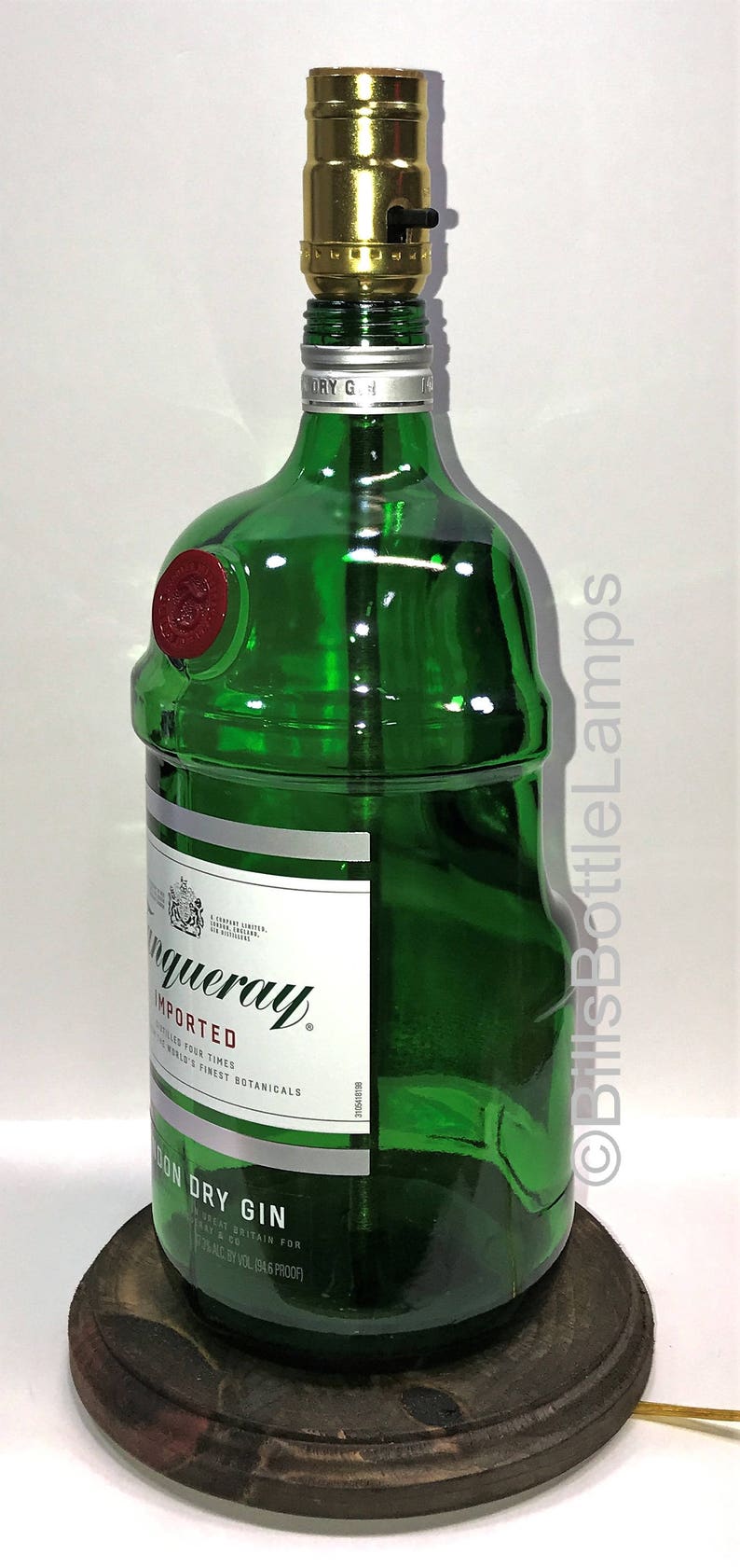 Puede incluir: Una botella de vidrio verde con una pantalla de l&aacute;mpara dorada en la parte superior. La botella est&aacute; etiquetada como "Tanqueray Imported London Dry Gin" y tiene un sello de cera rojo. La botella est&aacute; sentada sobre una base de madera.