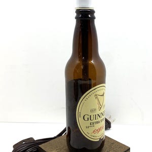 May include: A repurposed Guinness Extra Stout beer bottle transformed into a lamp. The brown glass bottle features a white label and a white lamp shade. It sits on a small, square wooden base with a brown cord and plug.