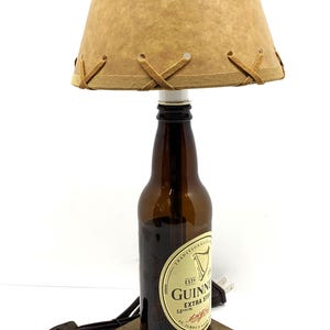 May include: A unique lamp crafted from a Guinness beer bottle, featuring a brown lampshade with decorative stitching. The lamp sits on a wooden base and includes a power cord. The lamp is approximately 12 inches tall.