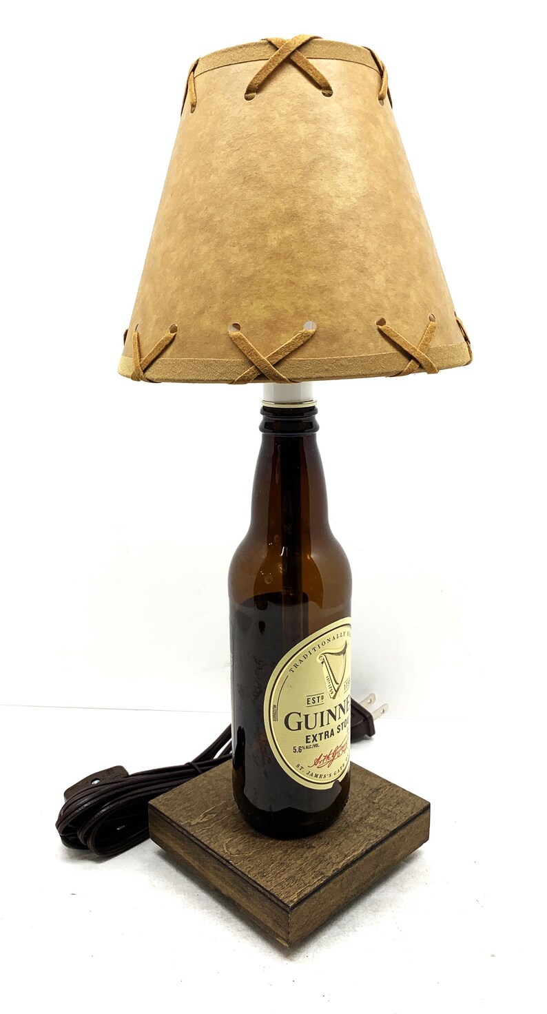 May include: A unique lamp crafted from a Guinness beer bottle, featuring a brown lampshade with decorative stitching. The lamp sits on a wooden base and includes a power cord. The lamp is approximately 12 inches tall.