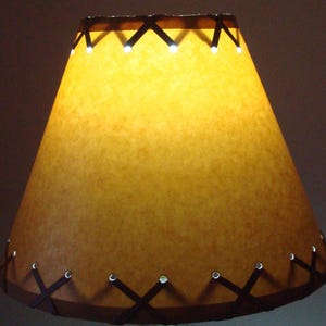 May include: A brown lampshade with a decorative stitched leather trim. The shade is made of parchment paper and has a warm, inviting glow.