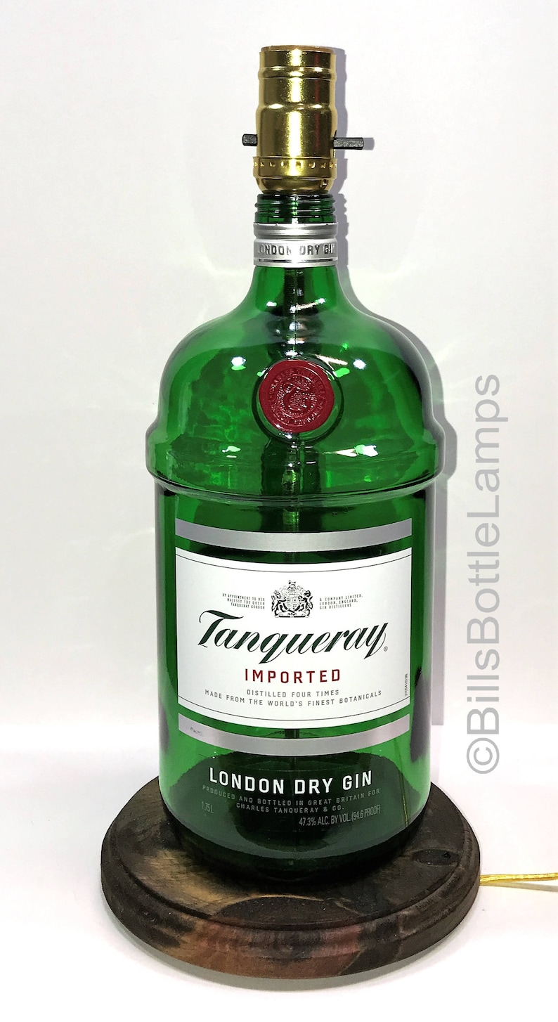 Puede incluir: Una l&aacute;mpara de botella de vidrio verde con una pantalla de metal dorado. La botella est&aacute; etiquetada con "Tanqueray Imported London Dry Gin" y tiene un sello rojo. La l&aacute;mpara est&aacute; sobre una base de madera.