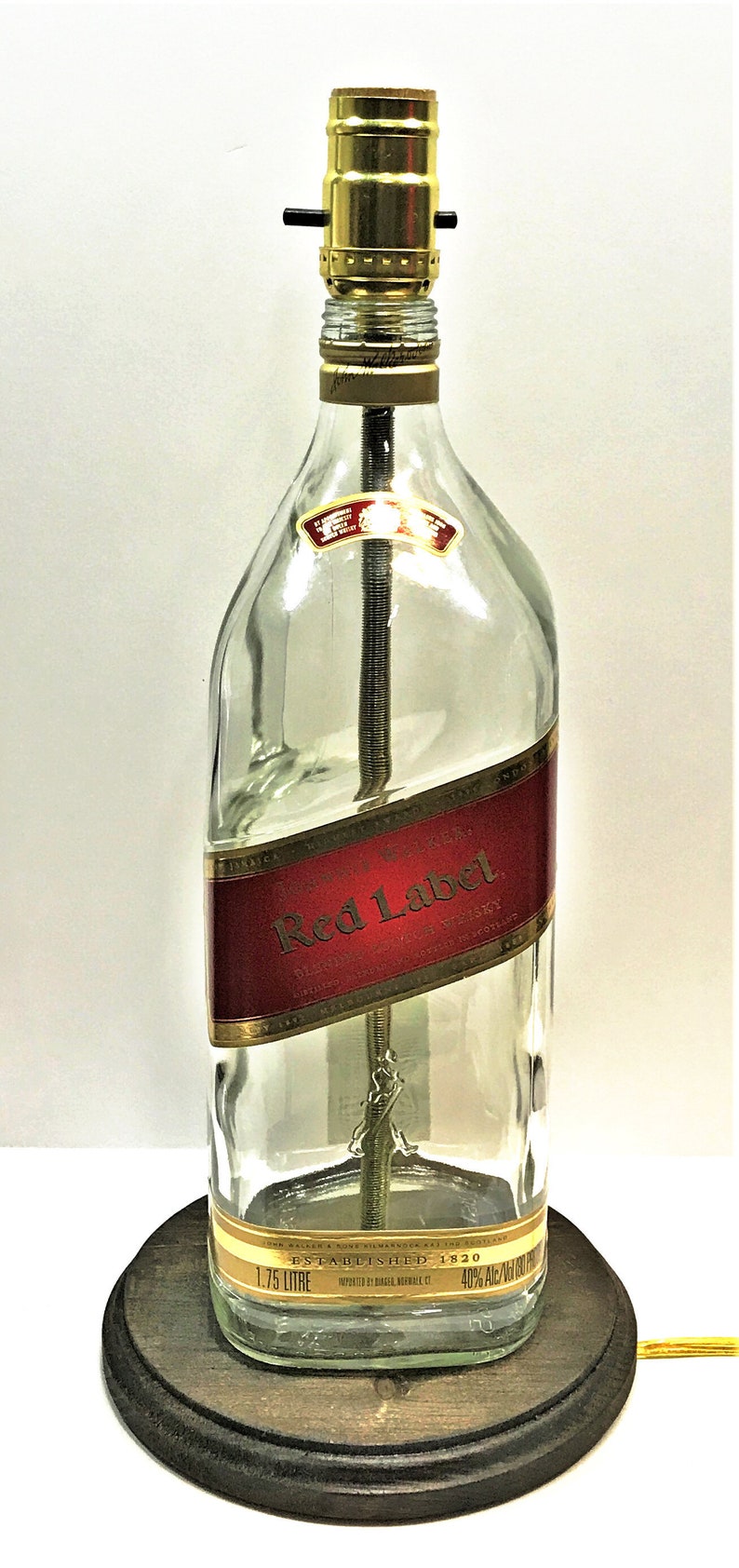 Puede incluir: Una l&aacute;mpara de mesa hecha de una botella de vidrio transparente con una etiqueta dorada que dice "Johnnie Walker Red Label Blended Scotch Whisky Established 1820 1.75 Litre 40% Alc./Vol.  Imported by Diageo North America, CT". La botella est&aacute; sobre una base de madera marr&oacute;n oscuro.