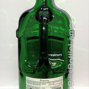 Puede incluir: Una botella de vidrio verde con asa, reconvertida en l&aacute;mpara. La botella est&aacute; etiquetada con "Tanqueray" y tiene una base de madera. La l&aacute;mpara tiene un casquillo de luz dorado.