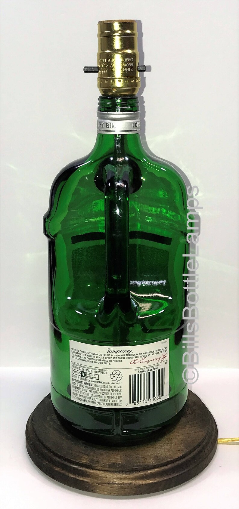 Puede incluir: Una botella de vidrio verde con asa, reconvertida en l&aacute;mpara. La botella est&aacute; etiquetada con "Tanqueray" y tiene una base de madera. La l&aacute;mpara tiene un casquillo de luz dorado.