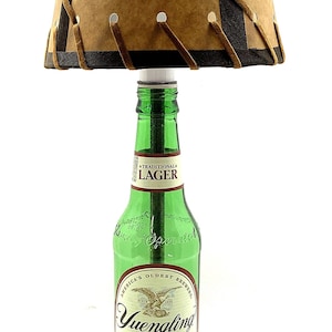 May include: A unique lamp made from a green beer bottle, featuring a tan lampshade with dark brown trim and lacing. The bottle is labeled "Yuengling Traditional Lager" and sits on a wooden base. The lamp includes a black power cord.